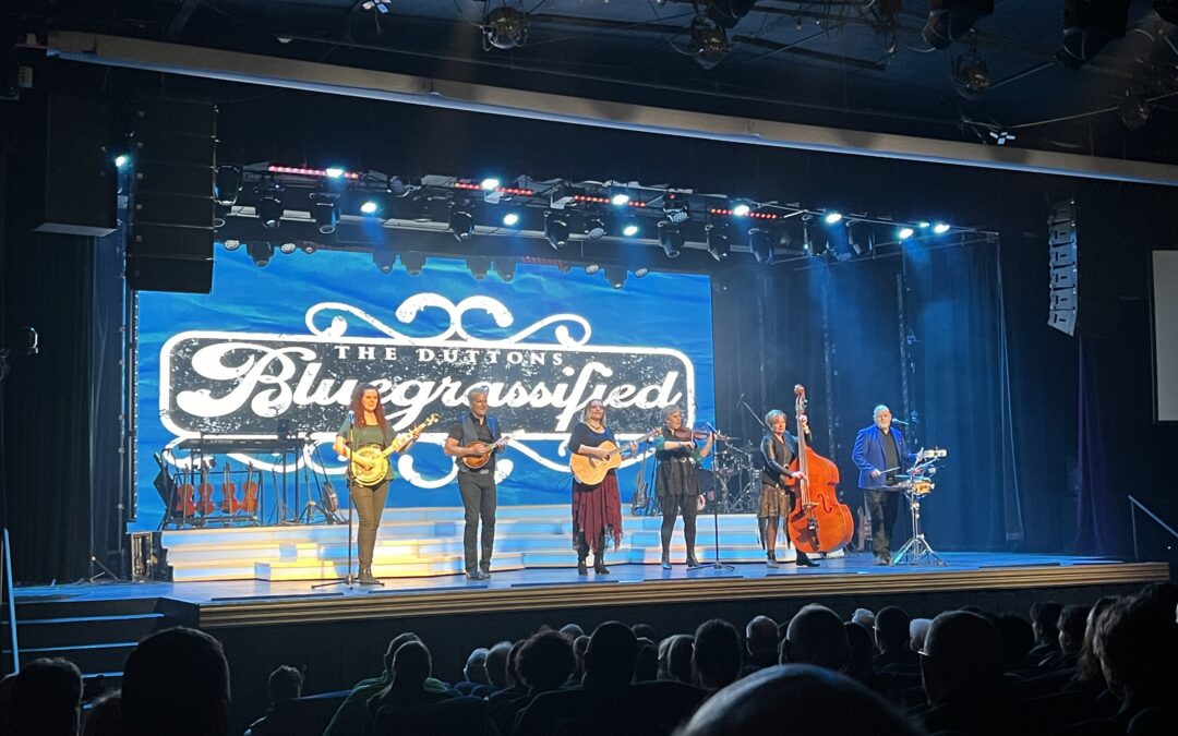 The Dutton family performing on stage at their 'Bluegrassified' show in Branson, MO, with members playing banjo, guitar, violin, and double bass against a backdrop with 'The Duttons Bluegrassified' written on it, under blue and white stage lighting